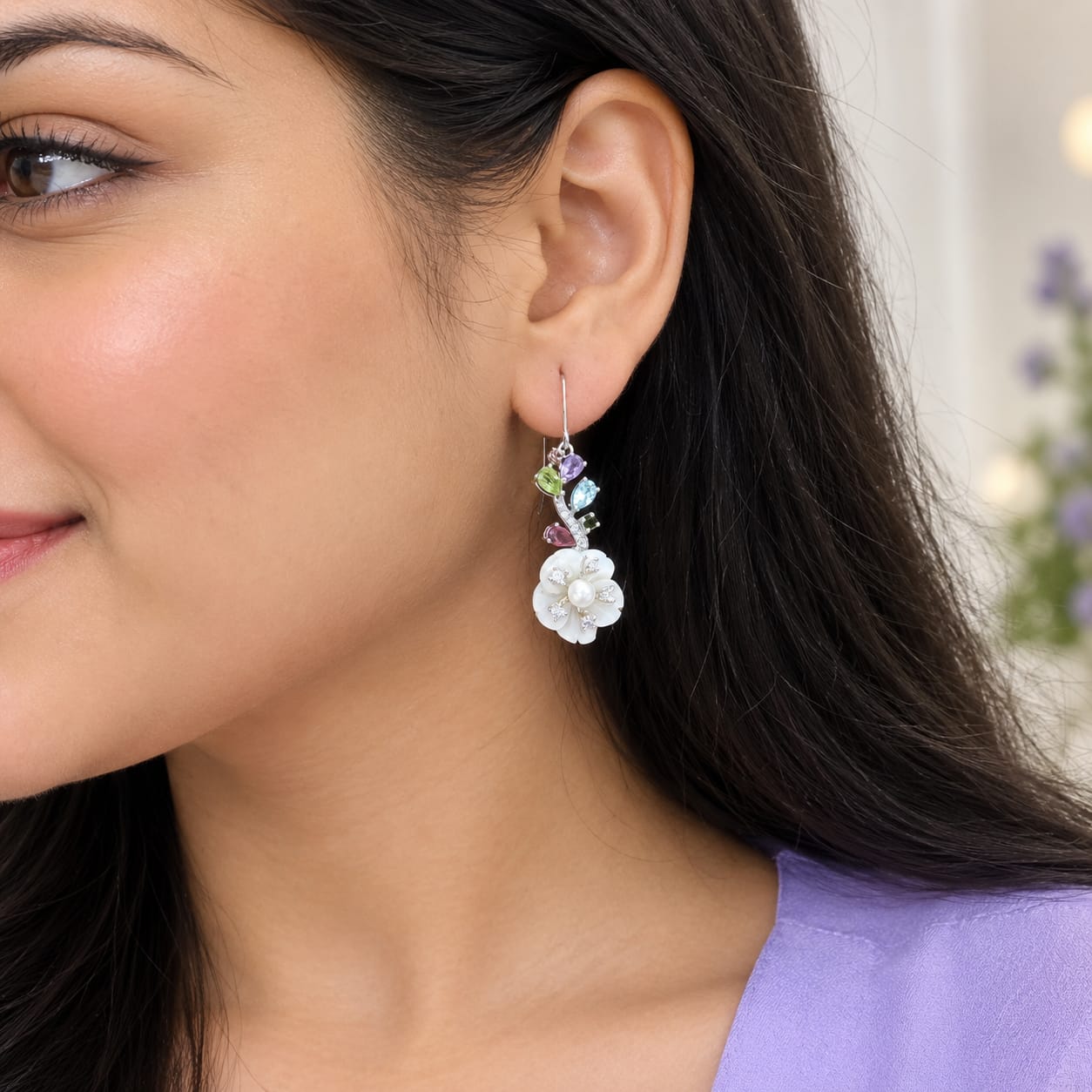 A close-up of a woman wearing a dangling earring with a white flower charm, small green and pink stones, and a silver-toned setting.