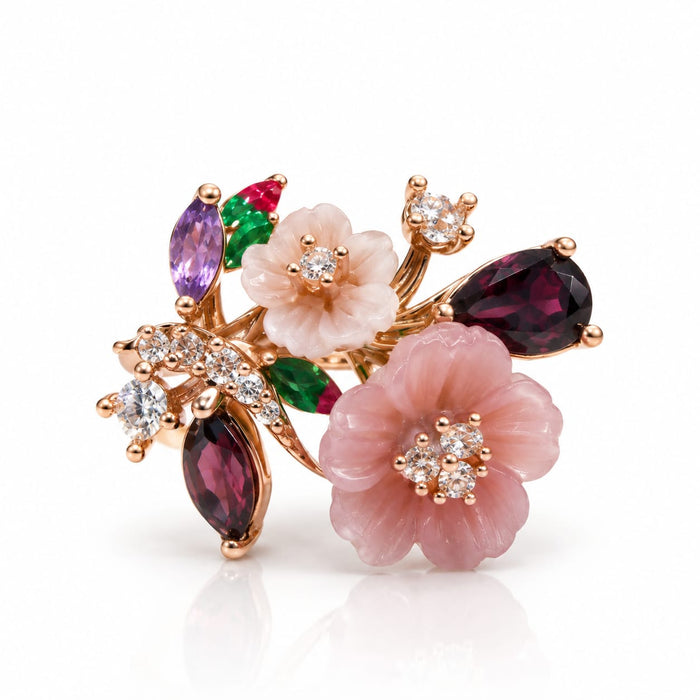 A floral rose gold brooch featuring two pink cabochon flowers, marquise-cut dark magenta garnet leaves, emerald-cut purple and green garnet accents, and small clear crystal clusters, shown on a white background with a soft reflection.