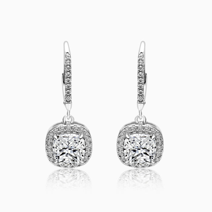 A pair of silver dangling grand solitaire earrings with a central cubic zirconia surrounded by smaller zirconia crystals, attached to silver hooks.