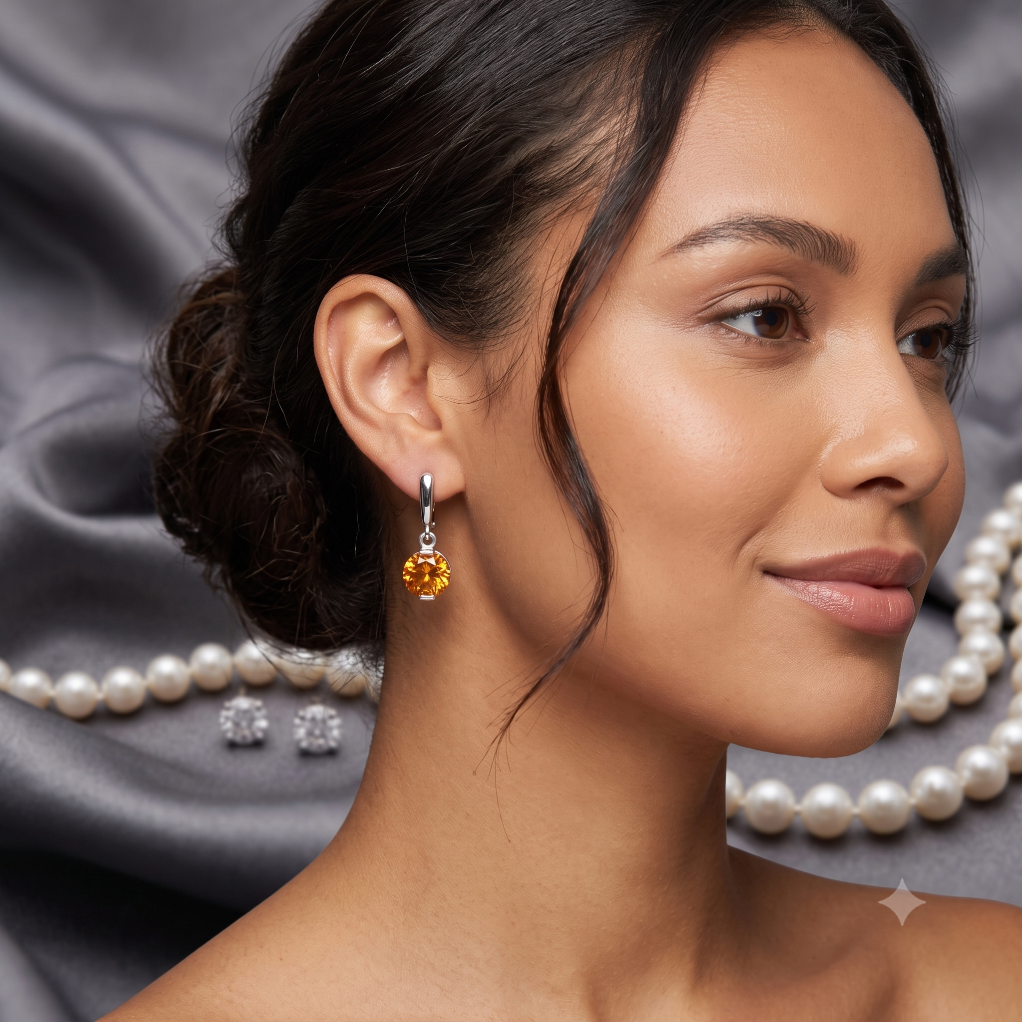 A close-up lifestyle shot of a woman wearing a silver-toned drop earring with a round yellow gemstone, set against a dark gray satin background with pearl strands.
