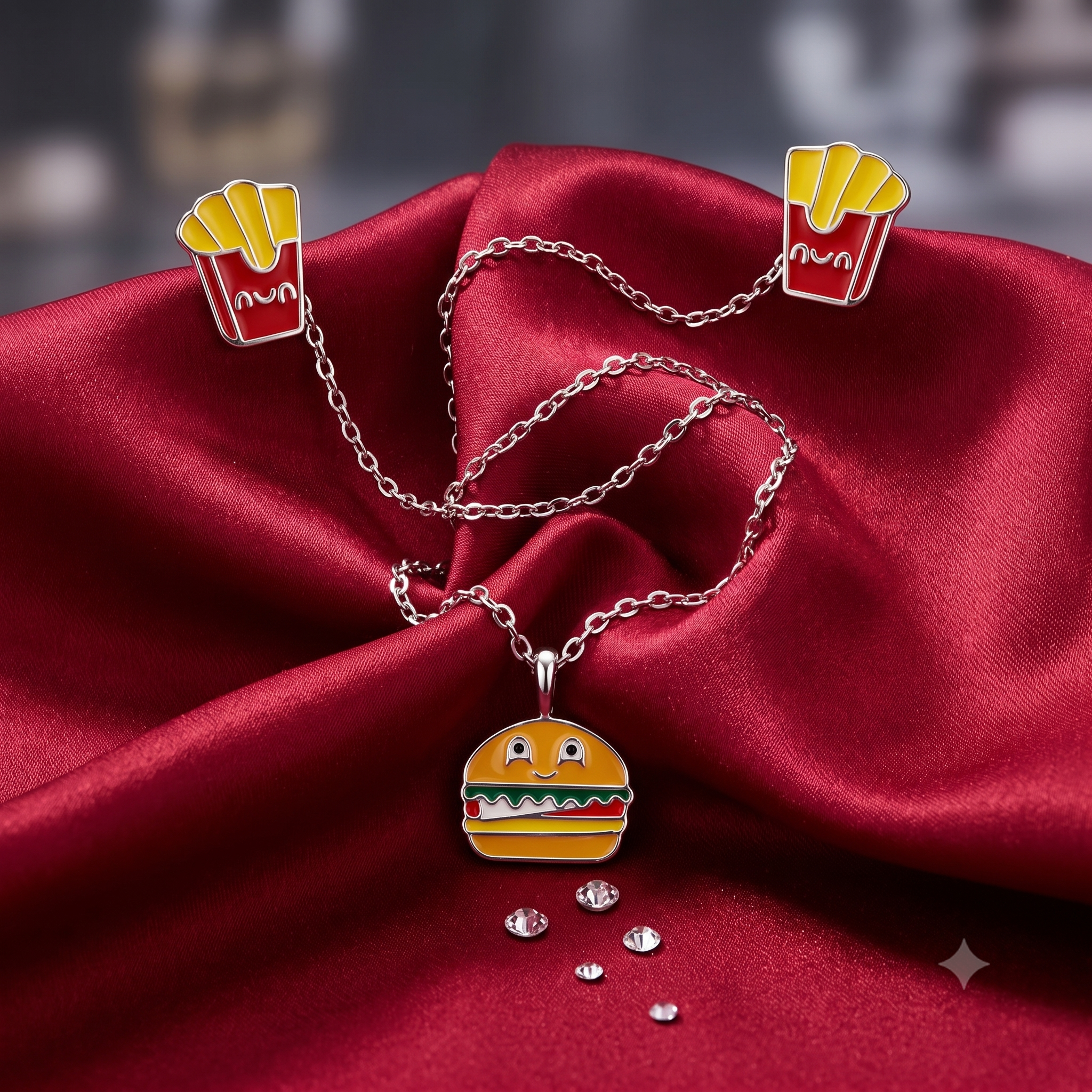 A silver-tone necklace with a cute cartoon hamburger pendant, displayed on draped red satin fabric. The necklace is shown with two matching hamburger-and-french-fries lapel pins and a few loose rhinestones.
