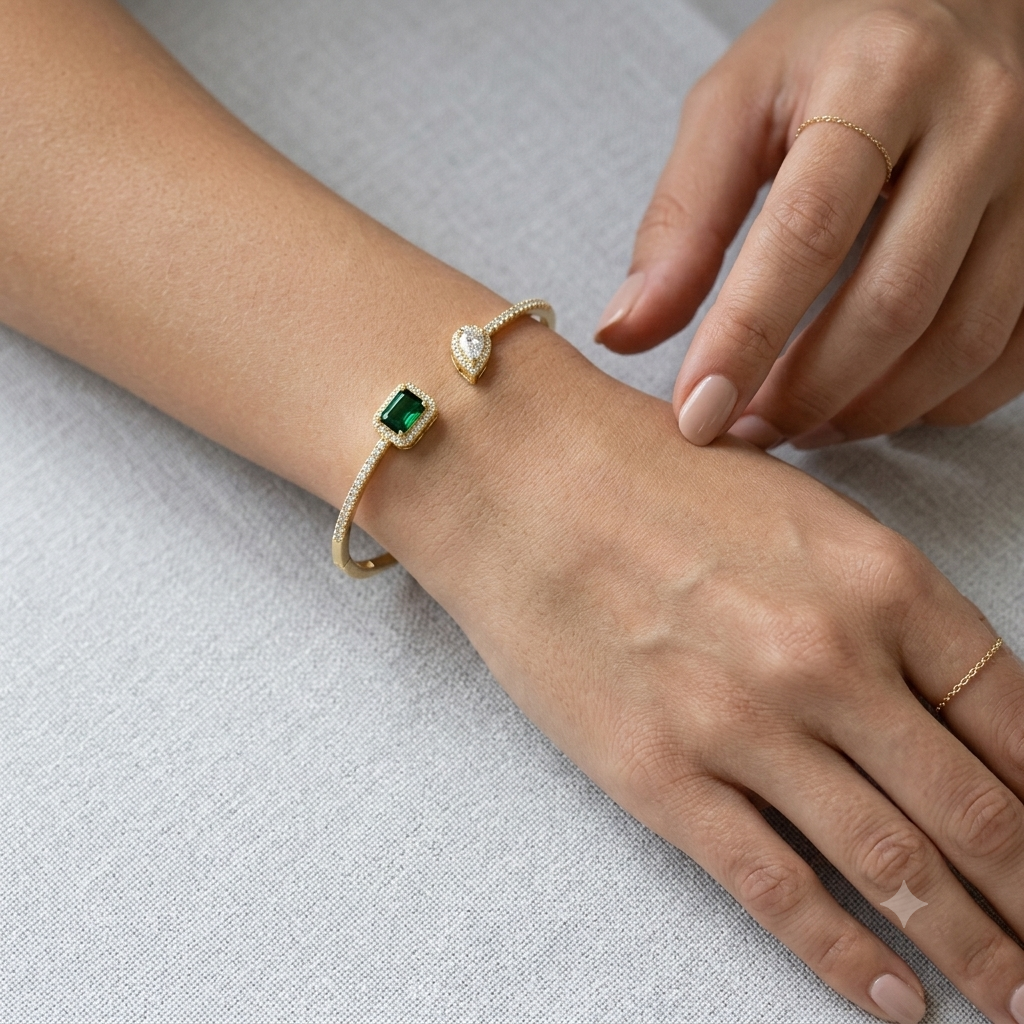 A gold open cuff bracelet with a rectangular green gemstone and a teardrop-shaped clear stone, shown worn on a person's wrist with two hands resting on a light gray textured surface.