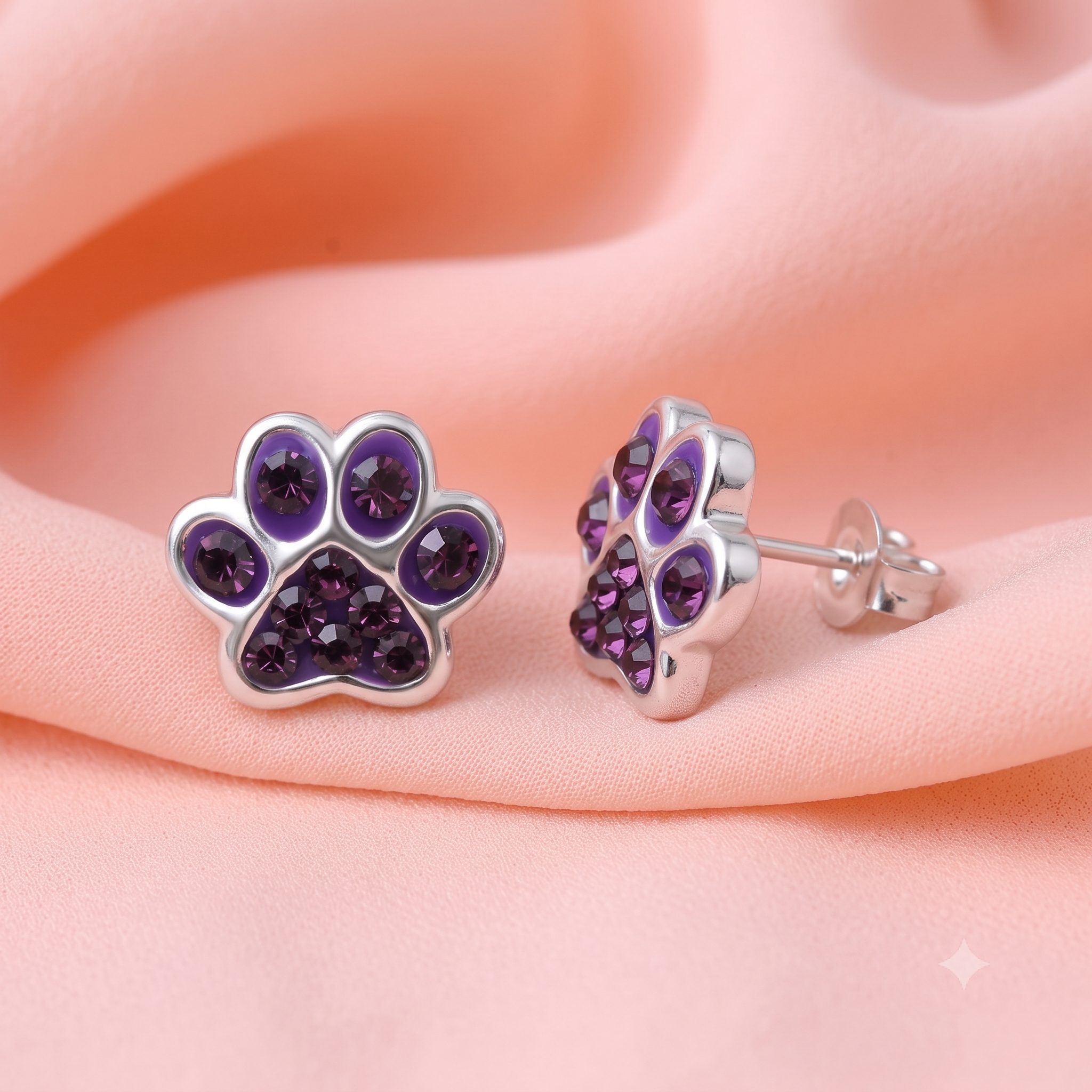 A pair of silver paw-shaped stud earrings set with deep purple crystals, displayed on a soft peach fabric background.