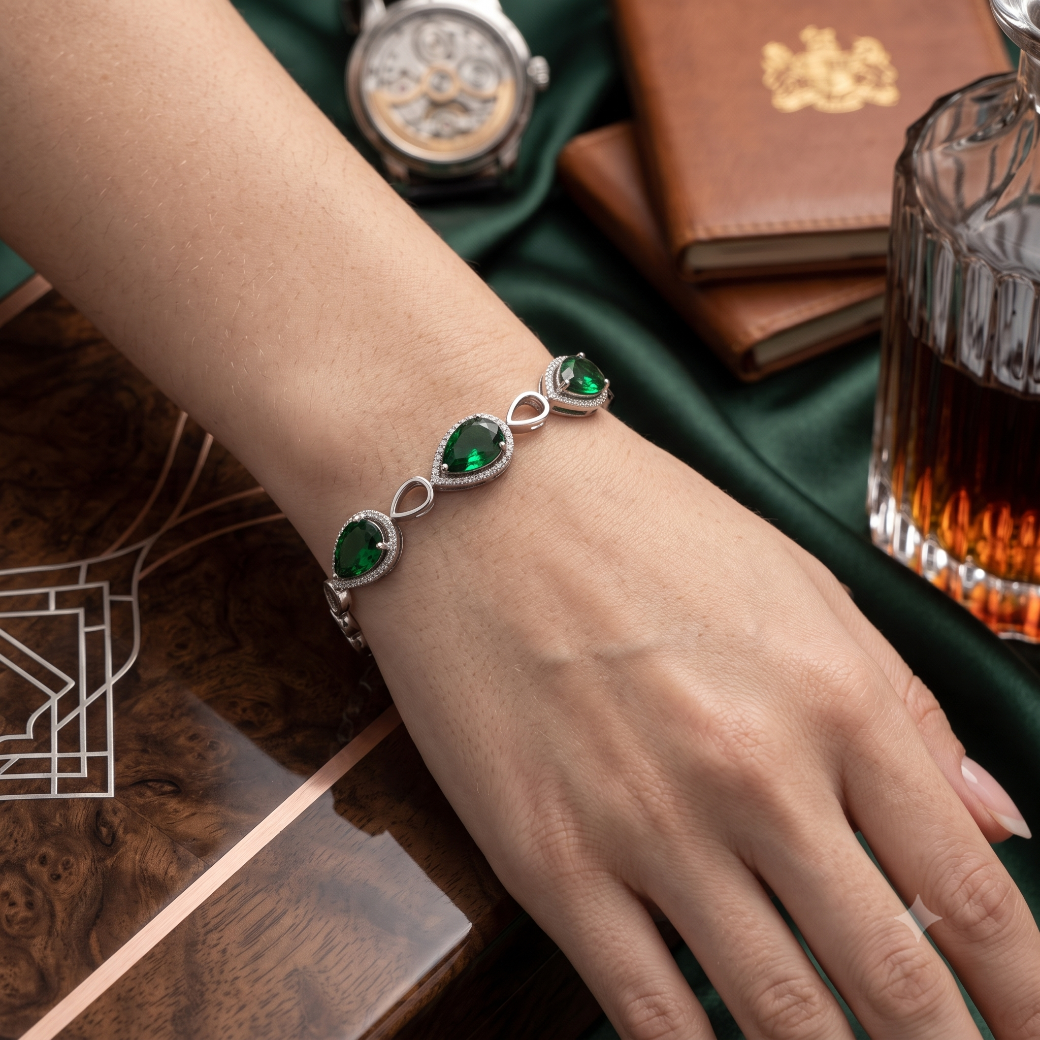 A close-up of a person's hand wearing a silver bracelet with three green teardrop-shaped gemstones set in silver, resting on a dark green satin cloth with a watch, leather notebook, and glass decanter nearby.