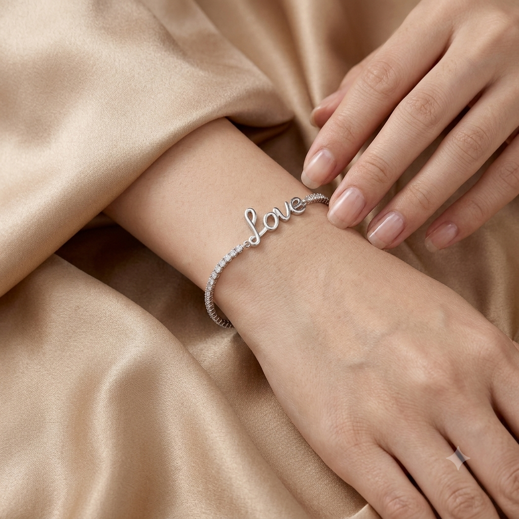 A delicate silver bracelet with the word 'love' in cursive script, embellished with small clear stones along one side, worn on a woman's wrist. The background consists of soft, draped champagne-colored satin fabric.
