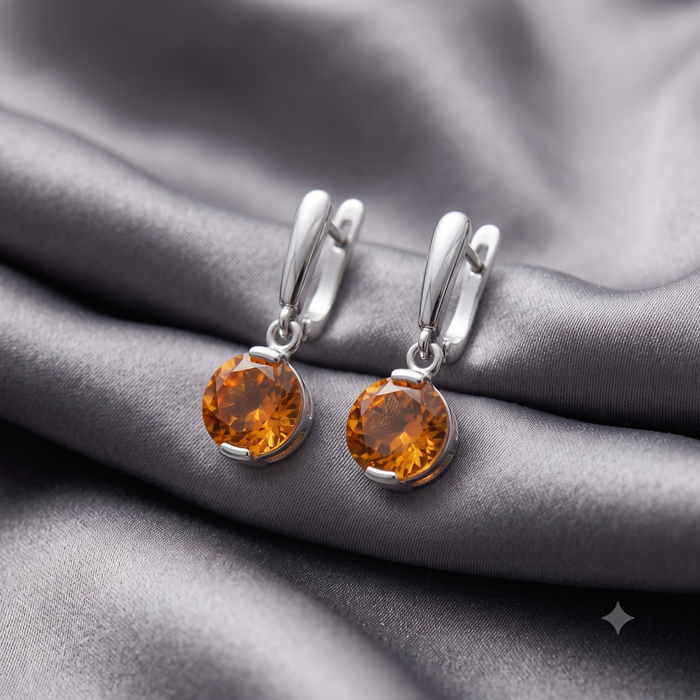A pair of silver-toned hoop earrings with faceted amber-orange gemstones, displayed on draped gray satin fabric.