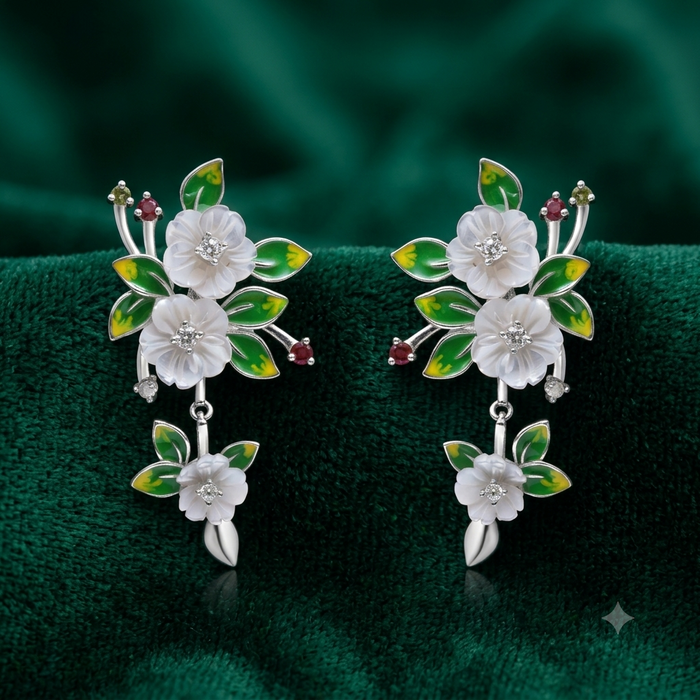 A pair of silver-toned dangle earrings decorated with white enamel flowers, green enamel leaves, and small red gemstone accents, displayed on a dark green velvet surface.