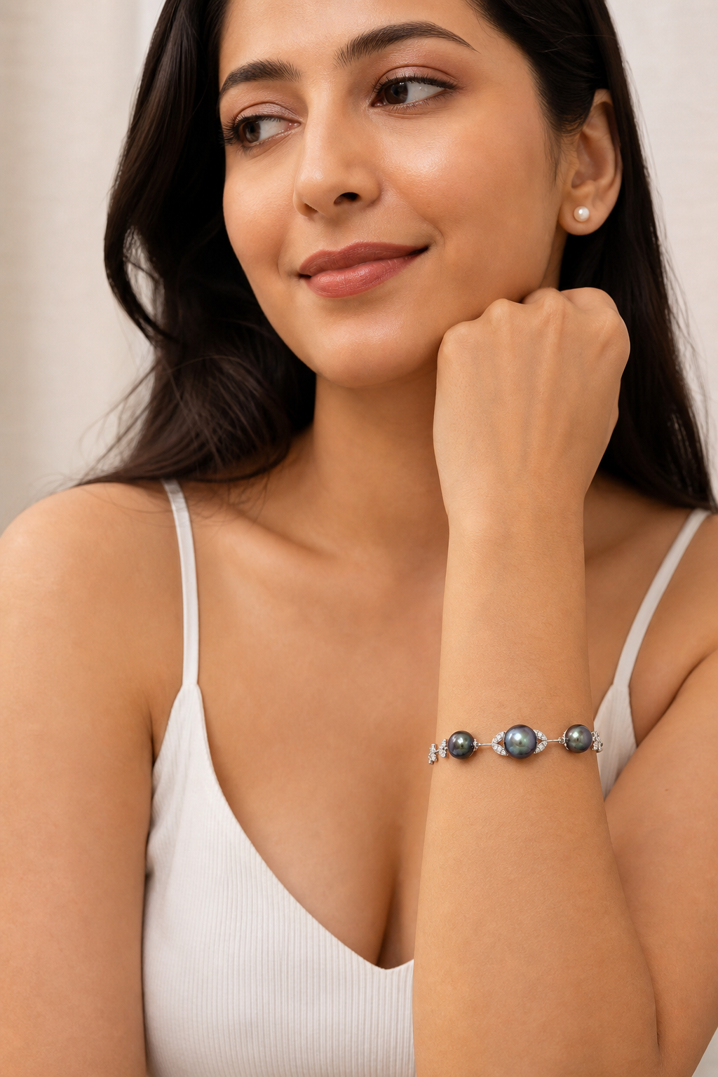 A woman wearing a delicate silver-toned bracelet with three dark grayish-blue round pearls, styled with a white ribbed camisole top.